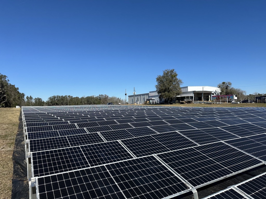 Ground-mount solar array in an open field beside a metal commercial building under a clear blue sky